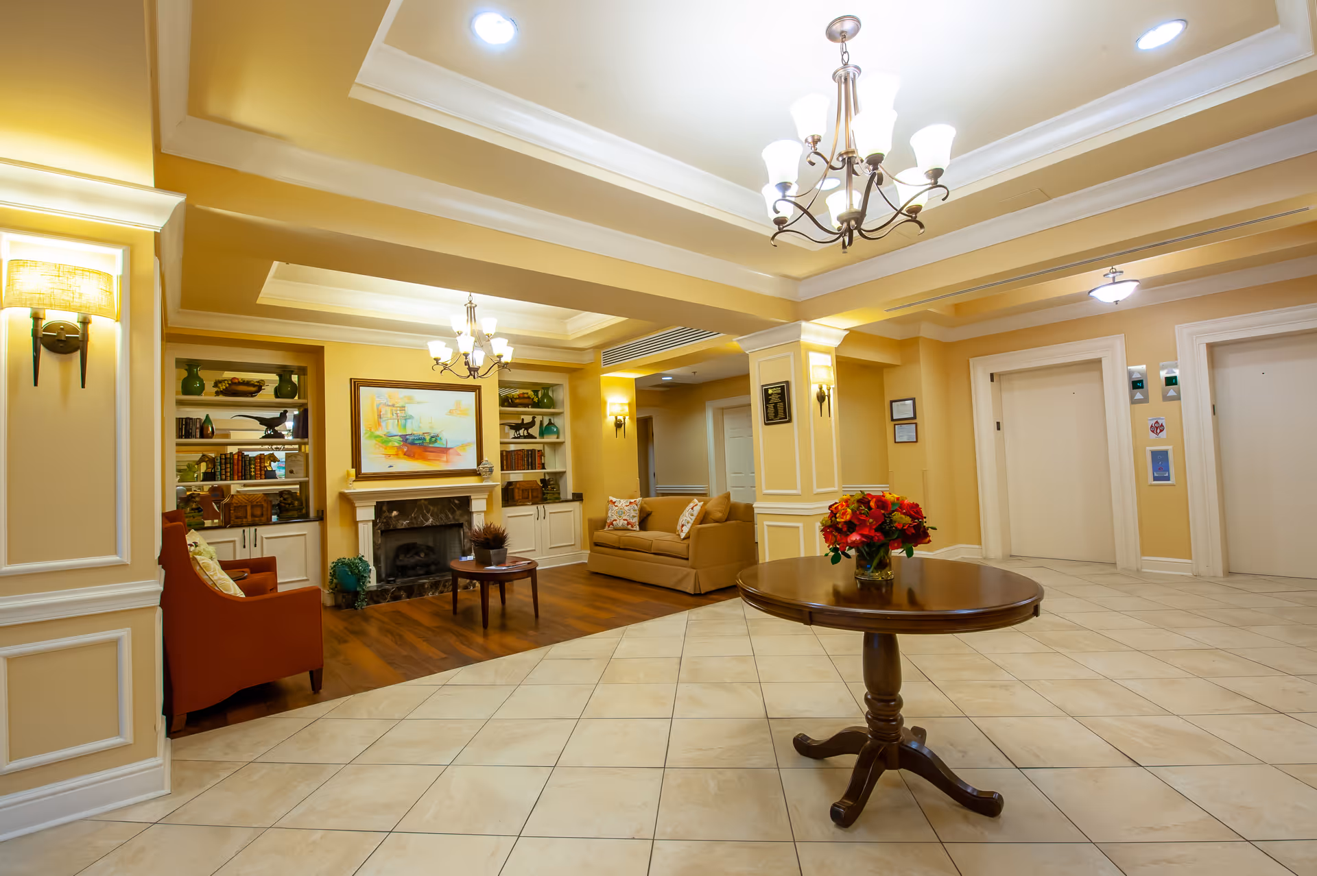 A spacious and well-lit senior living facility common area with beige walls and tiled floors. The room features a round wooden table with a vase of red flowers in the center, two elevators on the right side, and a cozy seating area with a sofa, armchair, coffee table, built-in bookshelves, and a fireplace with a colorful painting above it. Elegant chandeliers and wall sconces provide warm lighting.