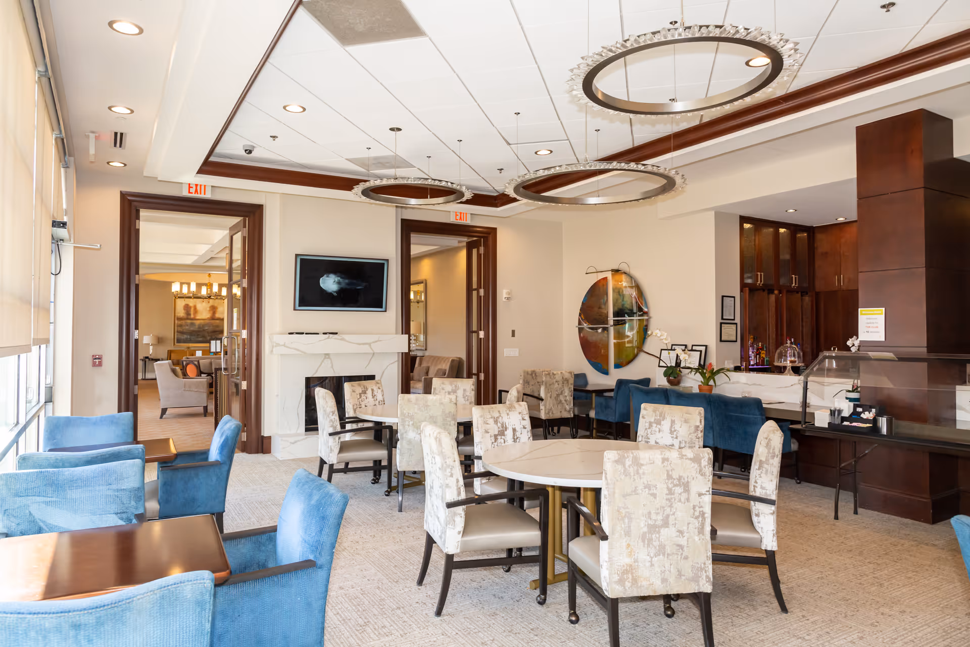 A bright and spacious dining area in a senior living facility with several round and square tables surrounded by upholstered chairs. The room features large windows with blinds, modern circular ceiling lights, a fireplace with a mounted TV above it, and a serving counter with dark wood cabinetry. There is colorful abstract wall art and a cozy seating area visible through open double doors.