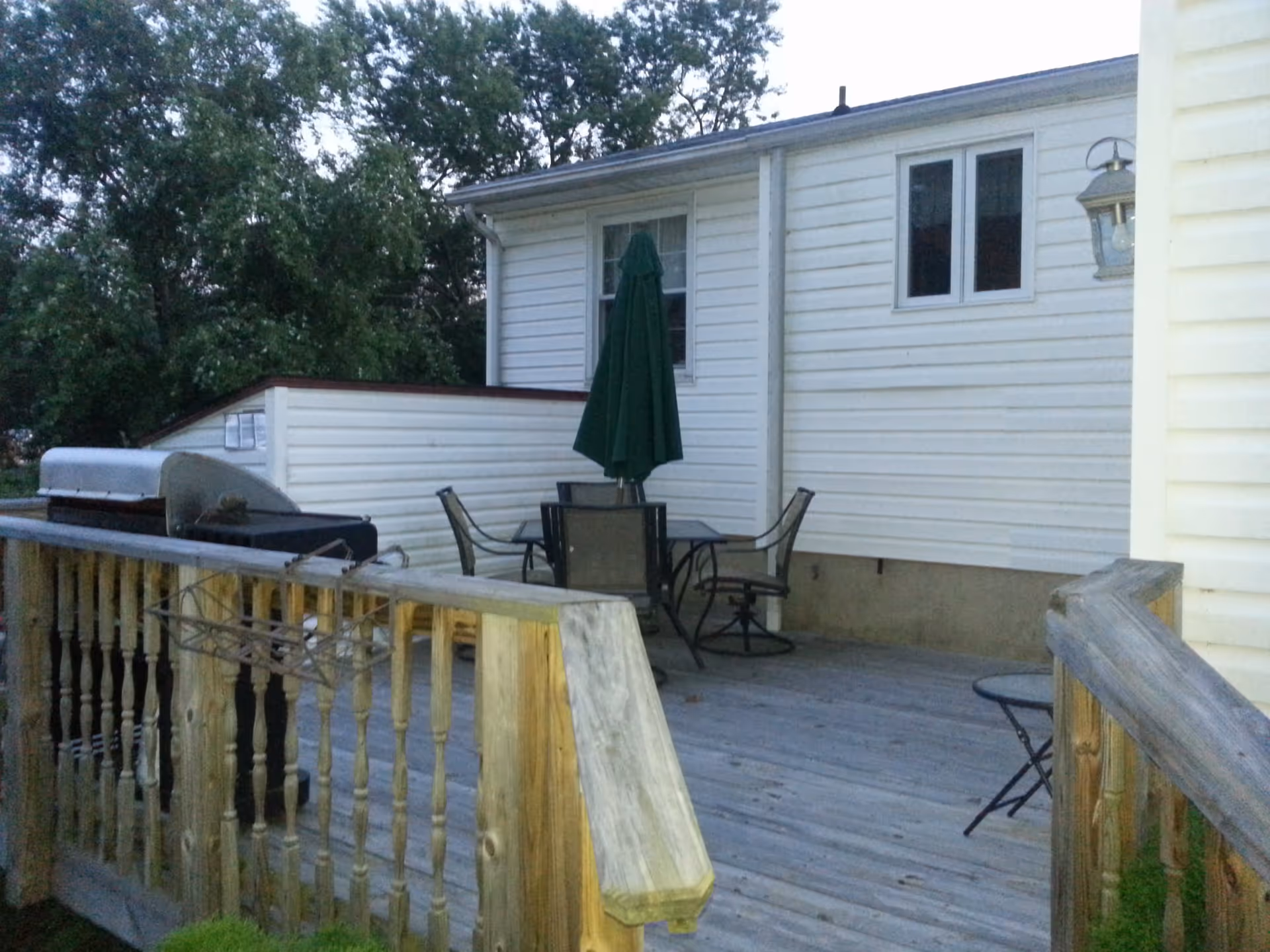 Outdoor wooden deck attached to a white house with a patio table, four chairs, a closed green umbrella, and a barbecue grill. Trees are visible in the background.
