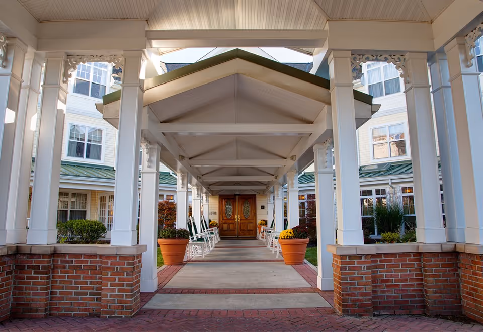Covered entrance walkway to a building with white pillars and brick bases, lined with white rocking chairs and large flower pots with yellow and red flowers, leading to double wooden doors with oval glass panels.