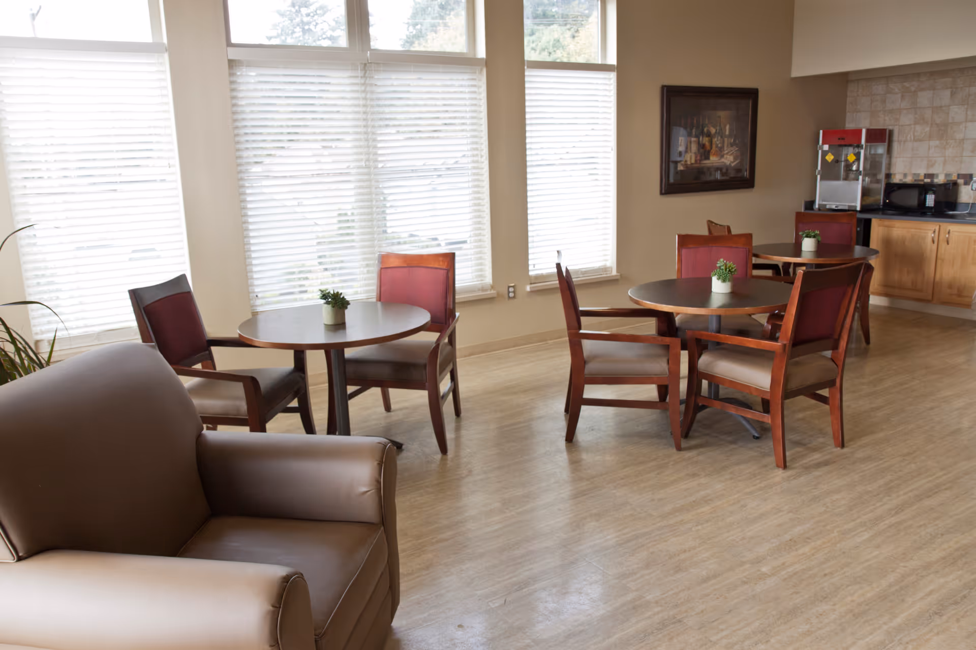 A bright room with large windows covered by white blinds, featuring round wooden tables with chairs around them. There is a comfortable armchair in the foreground, a popcorn machine and microwave on a countertop in the background, and small potted plants on the tables.