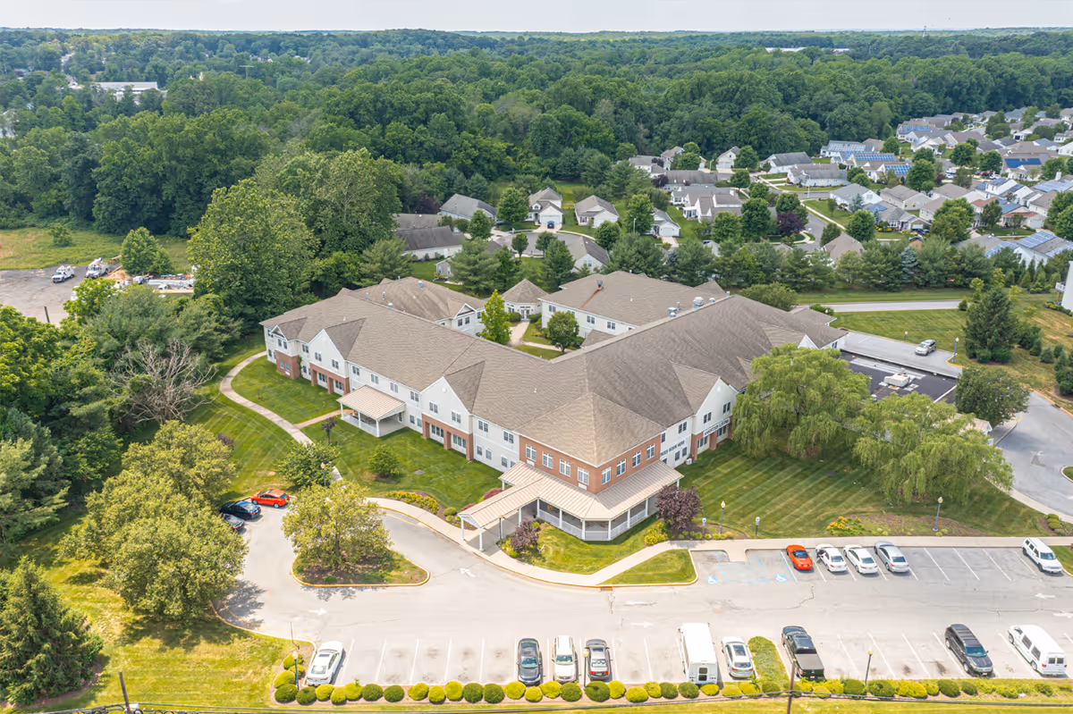 Aerial view of a large senior living facility building with parking lots, landscaped lawns, trees, and surrounding houses.