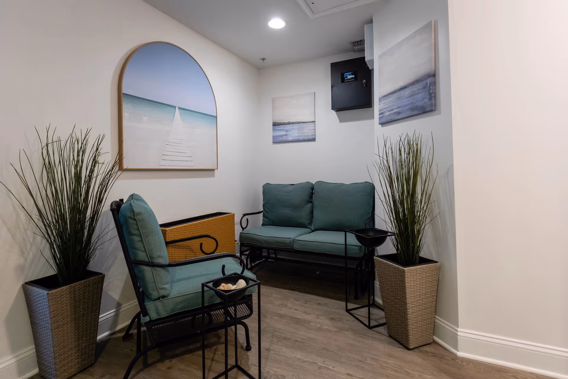 A small seating area with a cushioned loveseat and chair in teal color, two tall potted plants on either side, and three framed artworks on the white walls, including a large arched picture of a boardwalk leading to the ocean.