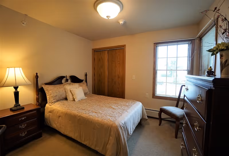A cozy bedroom with a double bed covered in beige bedding and pillows. There is a wooden nightstand with a lamp on the left side of the bed. A wooden dresser with decorative items and a chair are placed near a window with blinds. The room has beige walls, a ceiling light fixture, and a wooden closet door.