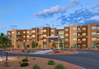 Exterior view of a modern multi-story senior living facility building with balconies, a covered entrance, and landscaped surroundings under a partly cloudy sky.