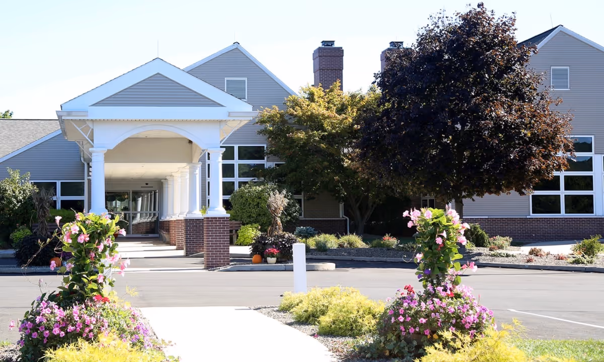 Front exterior view of The Meadows, A Park Home Personal Care Community, showing a covered entrance with white columns and brick bases, surrounded by landscaped flower beds and trees under a clear sky.