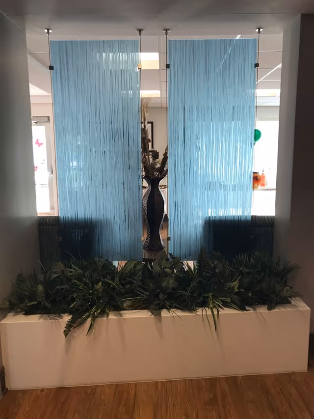 Indoor decorative area with a white rectangular planter box filled with green plants in front of two vertical blue string curtains hanging from the ceiling. Behind the curtains, a tall dark vase with dried floral arrangement is visible. The floor is wooden and the walls are light-colored.