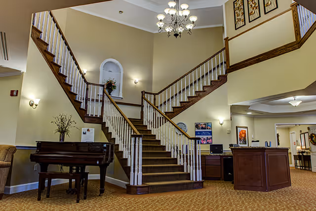 Interior view of a senior living facility lobby featuring a central carpeted staircase with wooden handrails and white balusters splitting into two directions at the top. To the left of the staircase is a grand piano with a vase of flowers on top. The walls are painted beige and decorated with framed artwork and wall sconces. To the right is a reception desk with office equipment and additional artwork on the walls. The ceiling has a chandelier and recessed lighting.