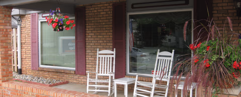 A covered outdoor porch area with two white wooden rocking chairs and a small white table between them. The porch has brick walls and columns, two large windows with closed blinds and maroon shutters, a hanging basket with red and purple flowers, and a planter with red flowers and tall grass-like plants.
