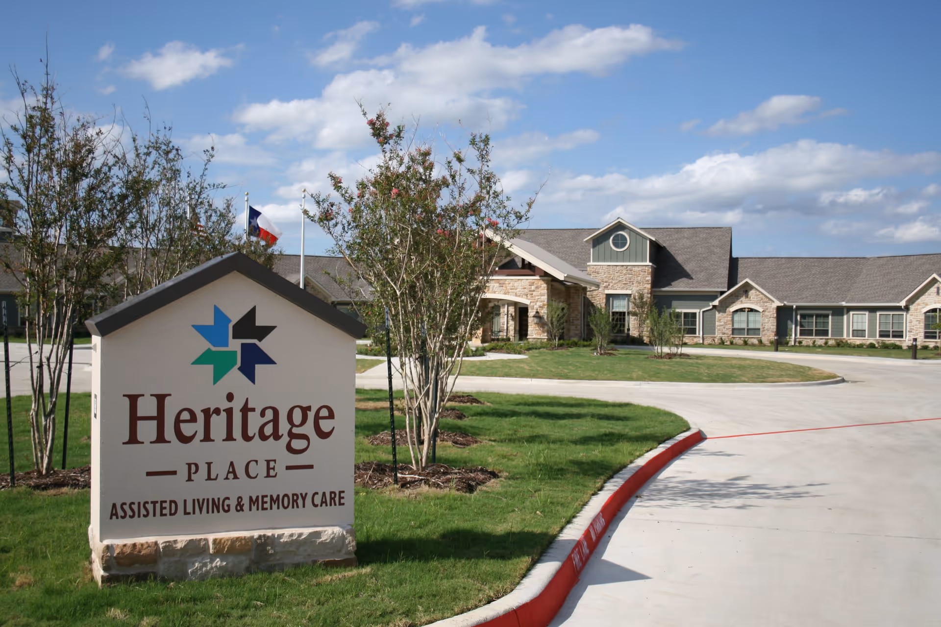 Exterior view of Heritage Place Assisted Living & Memory Care facility with a large sign in the foreground displaying the facility's name. The building is a single-story structure with stone and siding, surrounded by green grass, trees, and a curved driveway under a partly cloudy blue sky.