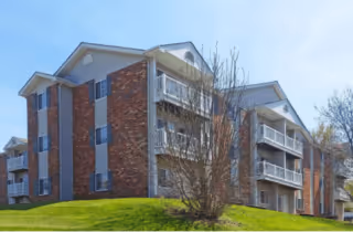 Exterior of a multi-story brick apartment building with balconies, a grassy lawn, and leafless trees under a blue sky.