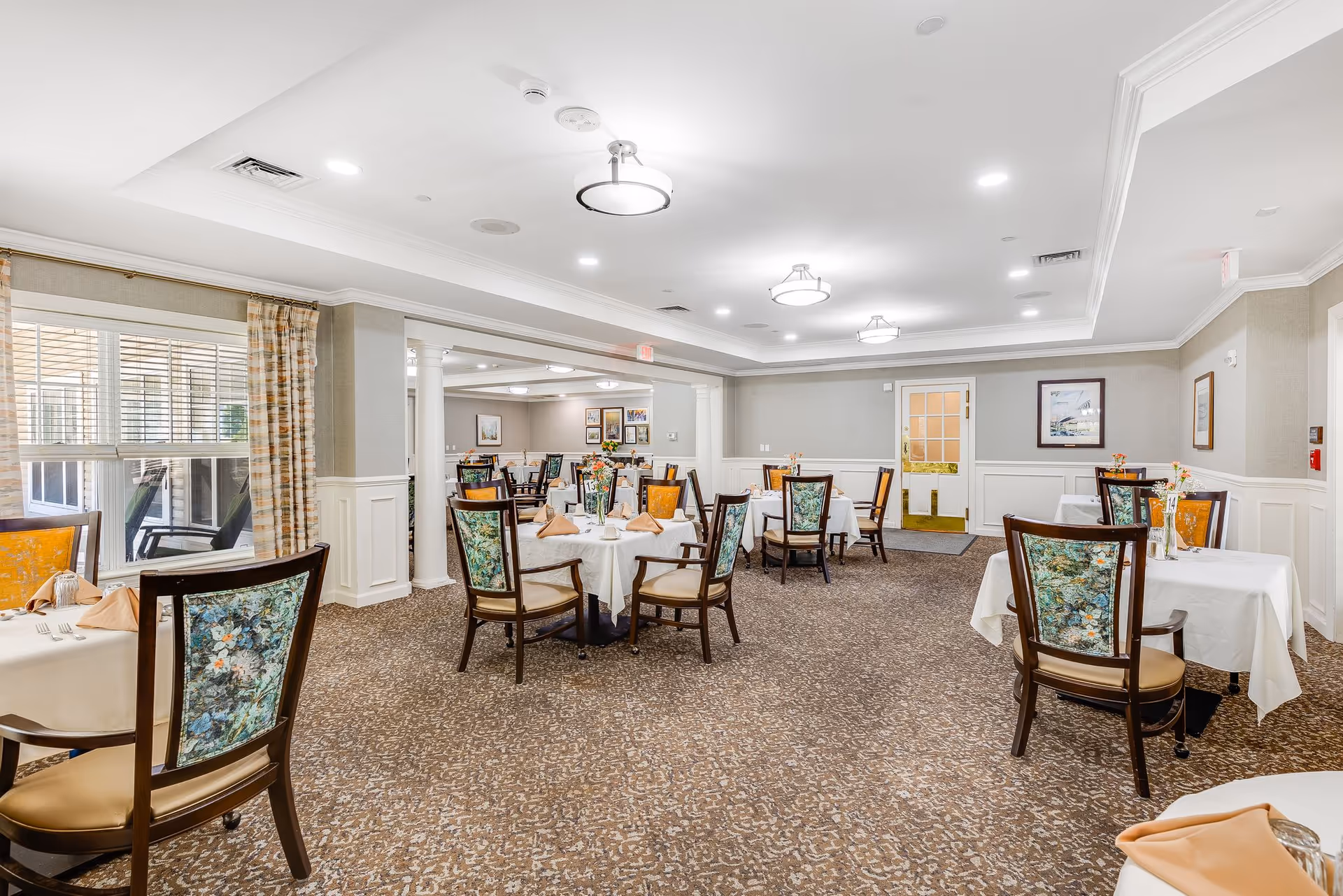 Bright, well-lit dining room with round tables set with white linens and floral-upholstered chairs.