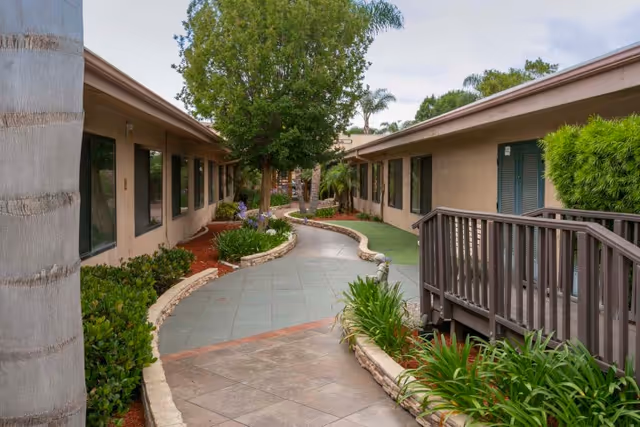 Outdoor walkway between two single-story buildings with beige walls and multiple windows, surrounded by landscaped greenery including bushes, trees, and plants. The walkway is paved and curves gently, with a small wooden ramp on the right side.