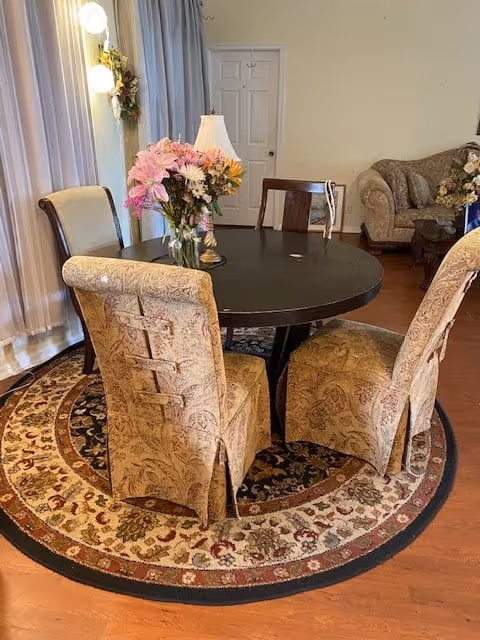 A cozy dining area with a round dark wooden table surrounded by four upholstered chairs with patterned fabric. A vase with pink and white flowers and a small lamp are placed on the table. The room has wooden flooring and a circular patterned rug underneath the table. In the background, there is a beige sofa with cushions and a side table with a floral arrangement. Light-colored curtains cover the windows on the left side.