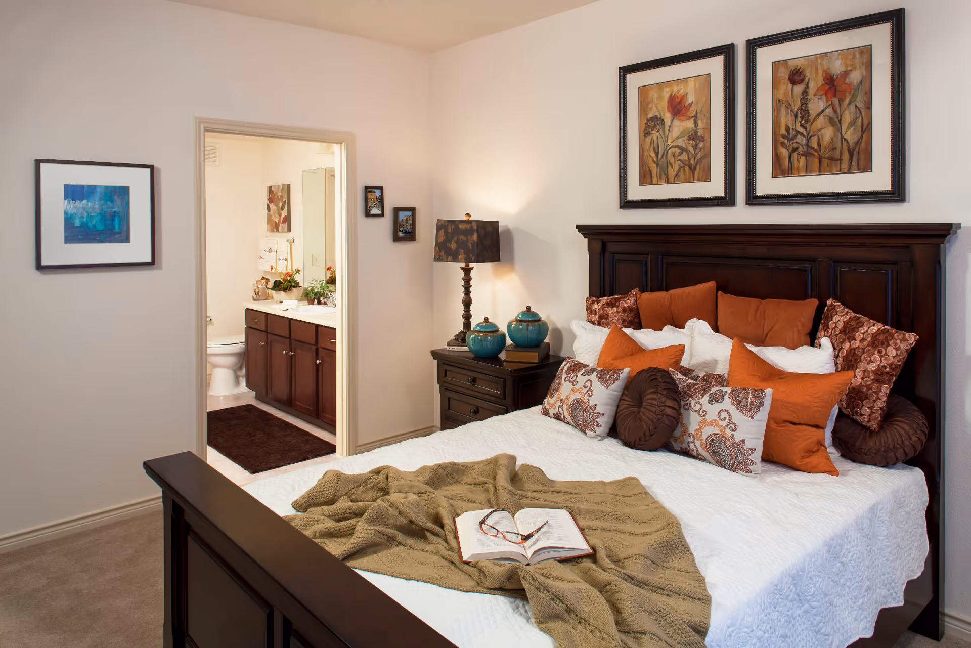A cozy bedroom with a dark wooden bed frame and a white bedspread adorned with multiple decorative pillows in shades of orange, brown, and white. A knitted beige blanket is spread across the bed with an open book and reading glasses on top. To the right of the bed is a dark wooden nightstand with a lamp and decorative teal jars. The wall above the bed features two framed floral paintings. An open doorway reveals a bathroom with a toilet, dark wooden cabinets, a white countertop, and a mirror. The bedroom walls are light-colored and there is a framed picture on the left wall.