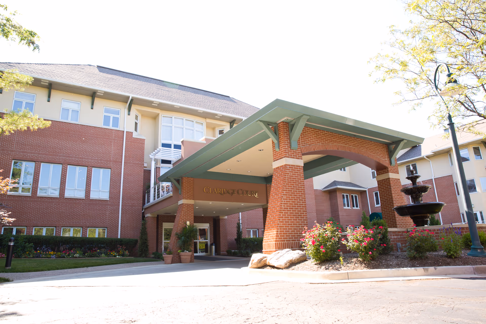 Front entrance of Claridge Court senior living building with a covered porte-cochere, brick facade, landscaping and a fountain.