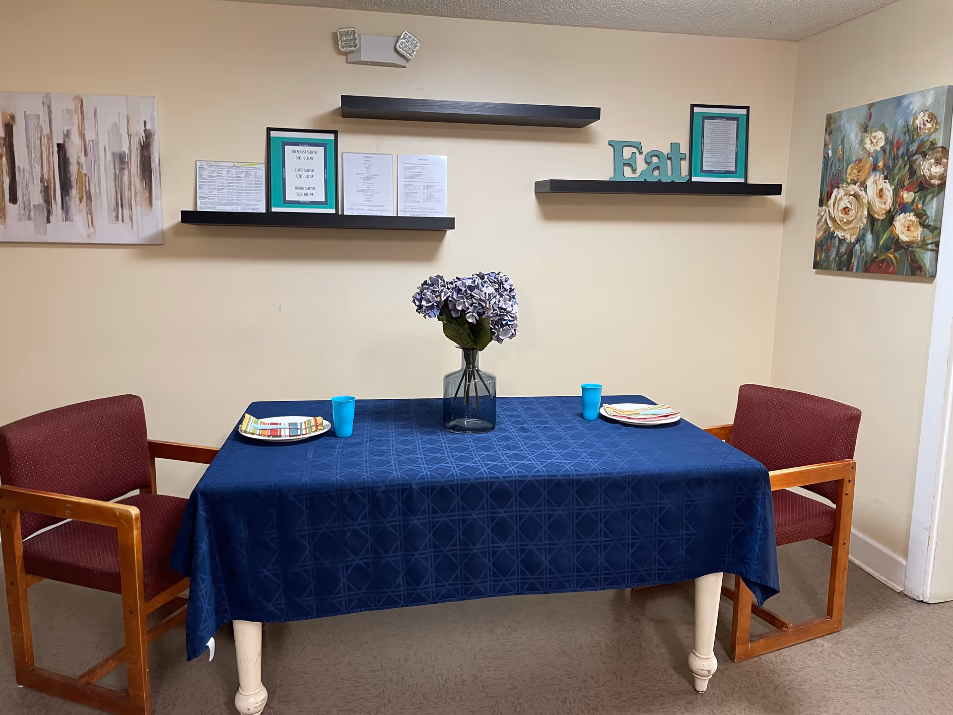 A small dining area with a rectangular table covered by a blue tablecloth, set with two plates, two blue cups, and a vase with purple flowers in the center. Two wooden chairs with red cushions are positioned on either side of the table. The beige walls have two floating black shelves holding framed documents and a decorative 'Eat' sign. Two paintings hang on the walls, one abstract and one floral.