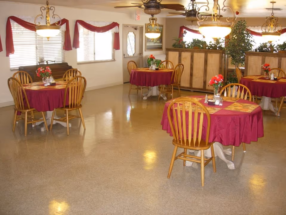 A dining room with several round tables covered with red tablecloths and wooden chairs around them. Each table has a small flower arrangement and placemats. The room has large windows with red valances, ceiling fans with lights, and a partition with plants on top.