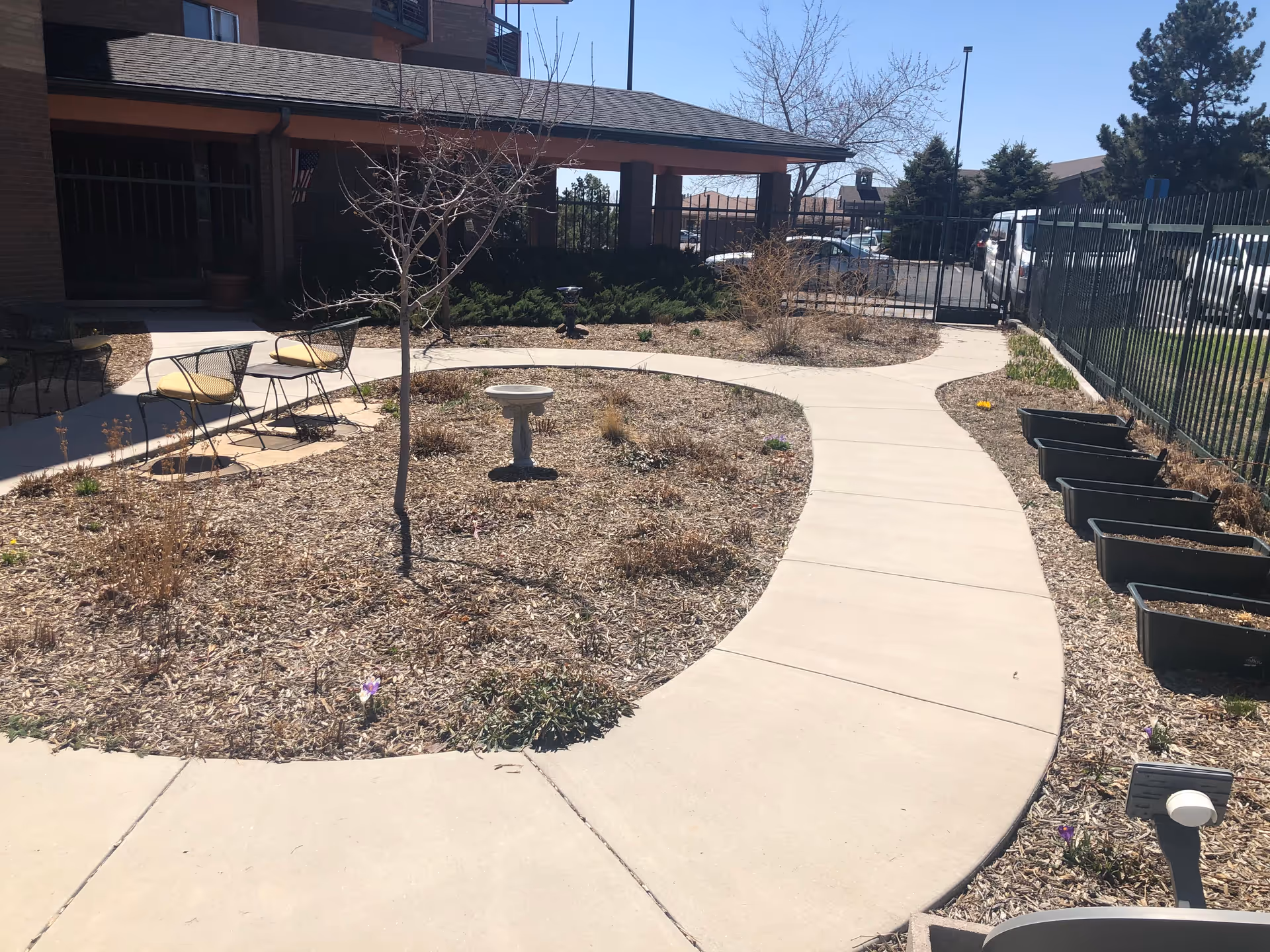 Curved concrete walkway through a landscaped courtyard with planters, a small birdbath, seating, and the building entrance in the background.
