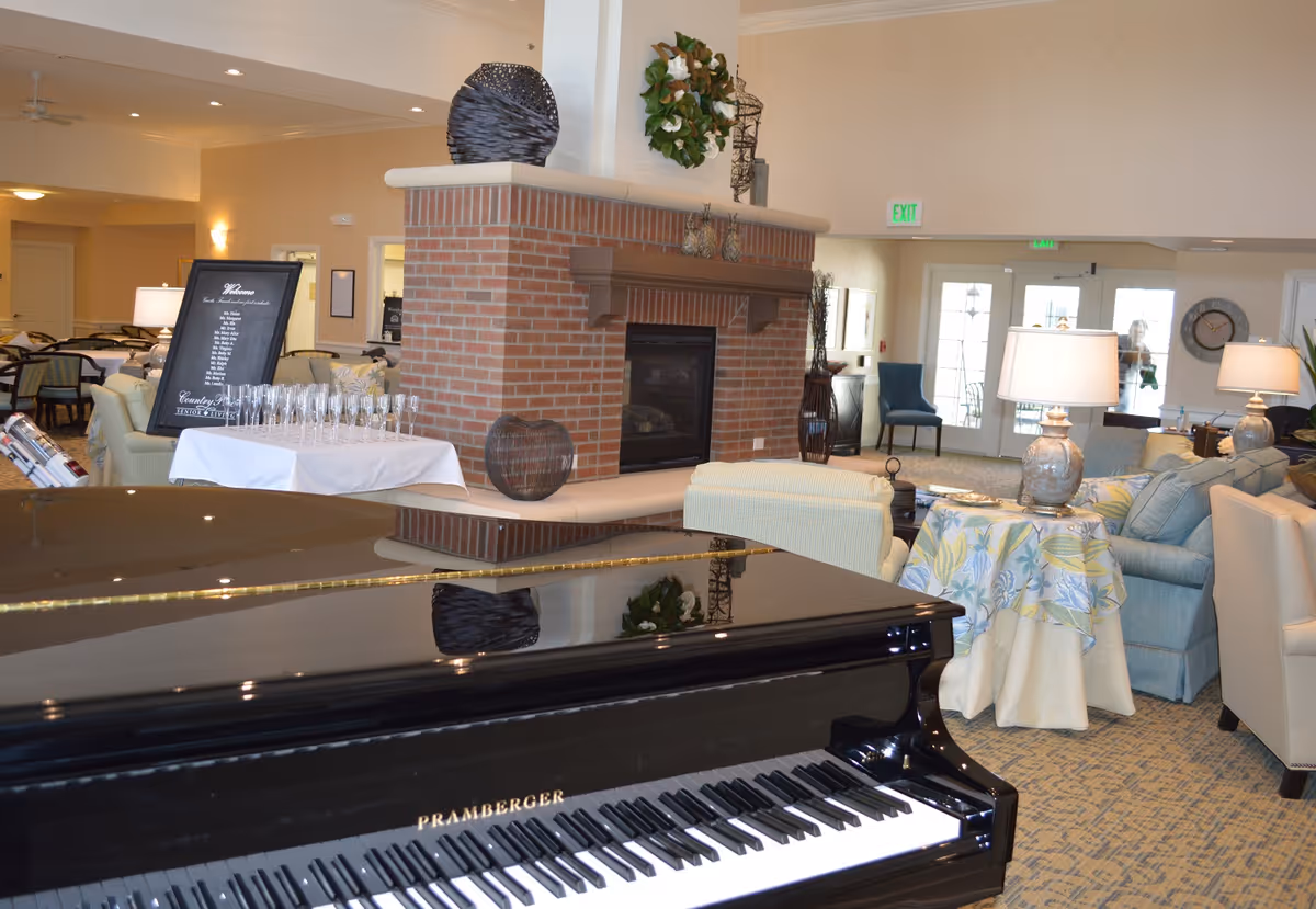 Interior view of a senior living facility common area featuring a black grand piano in the foreground, a brick fireplace in the center, and comfortable seating with sofas and armchairs arranged around tables with lamps and decorative items. There is a table with empty glasses and a framed menu or sign near the fireplace. The room has a warm, inviting atmosphere with neutral tones and soft lighting.