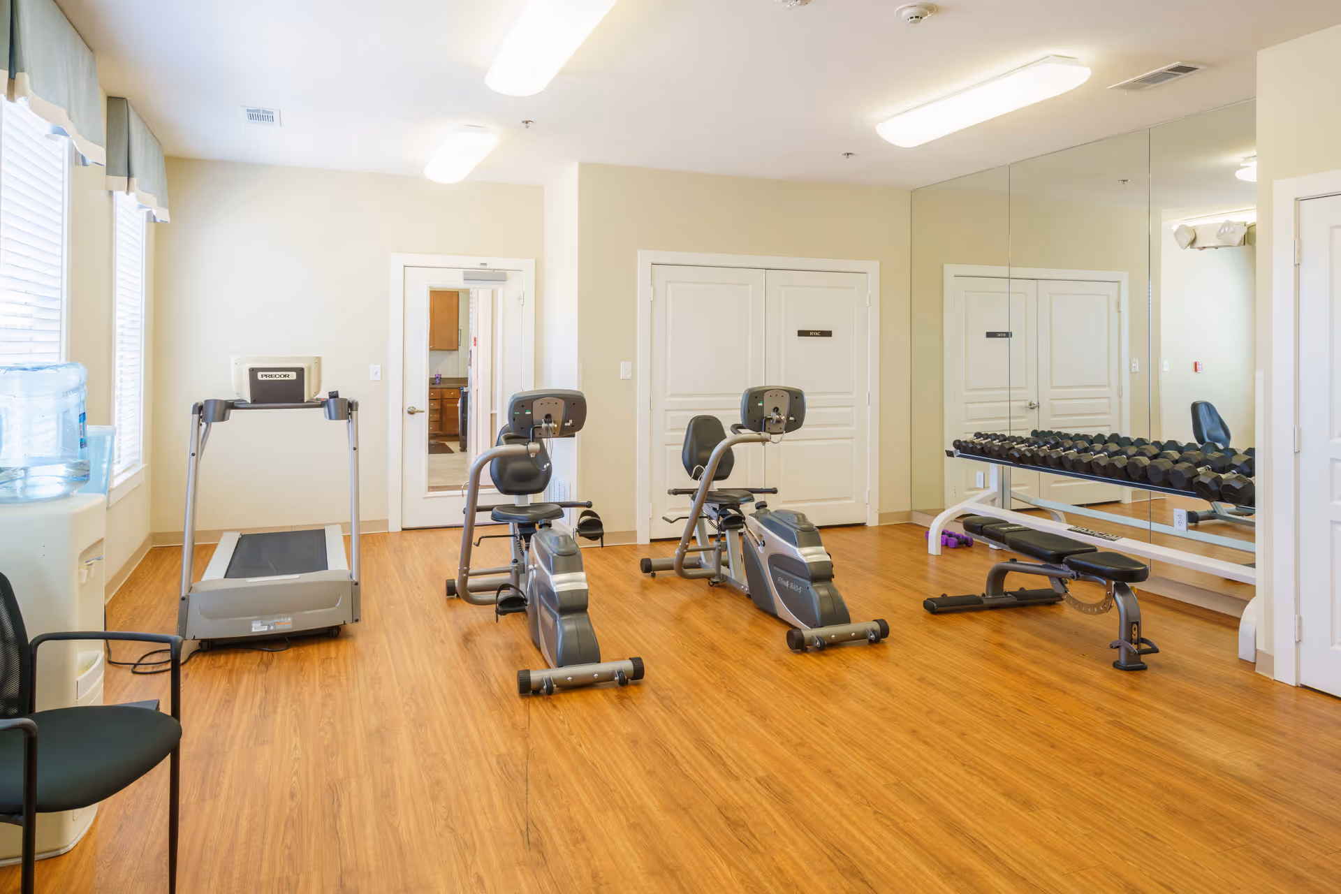 A small fitness room with wooden flooring featuring two recumbent exercise bikes, a treadmill, a bench, and a rack of dumbbells against a mirrored wall. There is a water cooler and a black chair near the windows on the left side, and two closed white doors on the far wall.