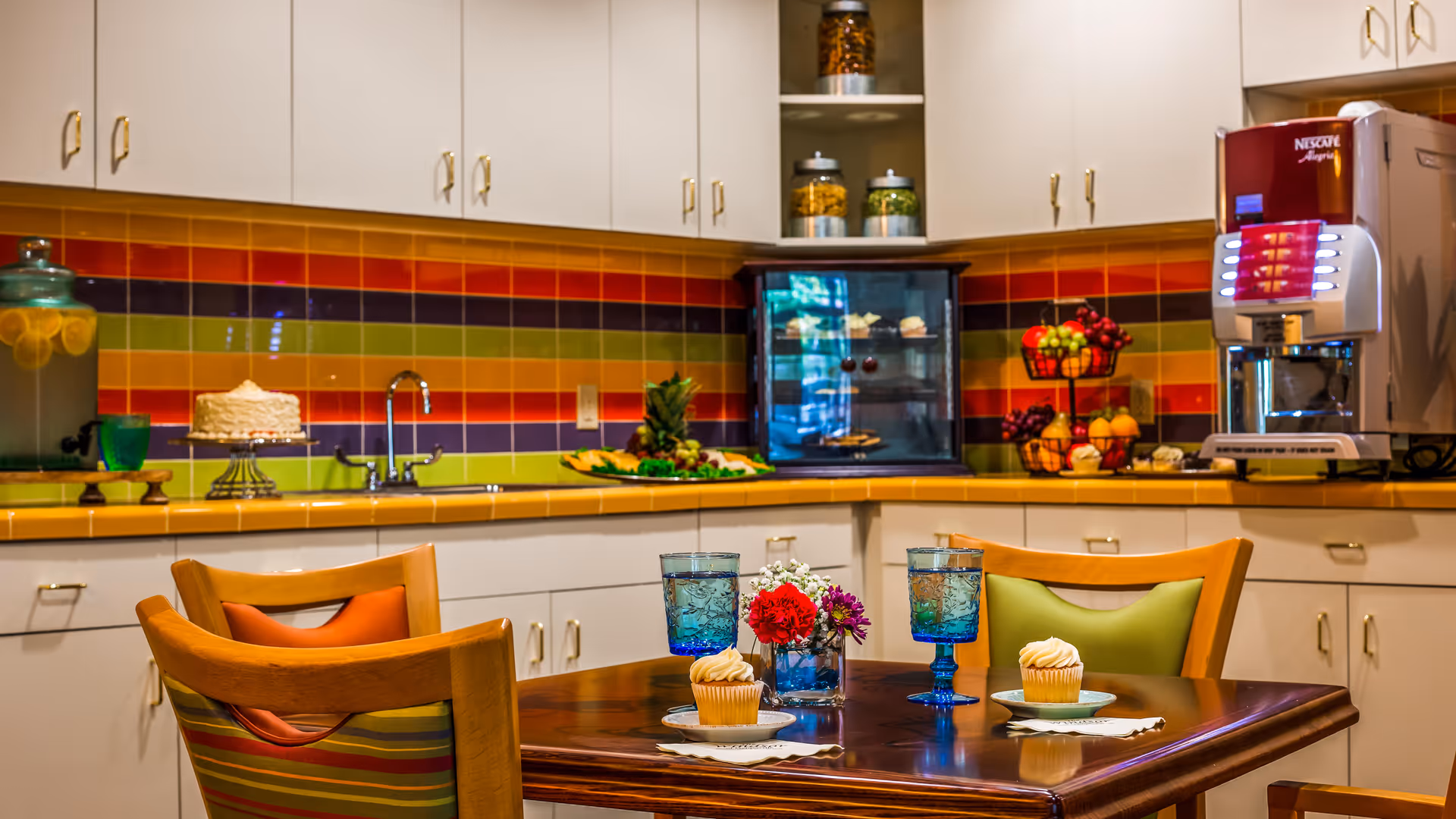 A colorful kitchen area with a wooden table set for two, featuring blue glasses, cupcakes on plates, and a small flower arrangement. The background shows white cabinets with gold handles, a multicolored striped tile backsplash, a sink, a cake on a stand, a fruit basket, a small display case with pastries, and a coffee machine.