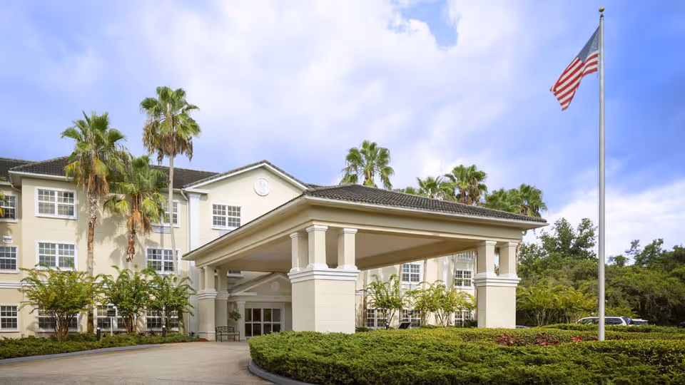 Exterior view of Colliers Assisted Living at Countryside, showing a three-story beige building with multiple windows, palm trees, a covered entrance with columns, and an American flag on a flagpole in front.