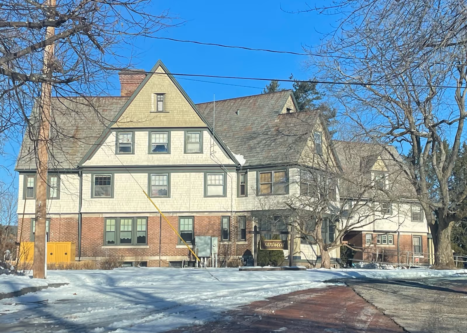 Large multi-story building with a steep roof and multiple windows, surrounded by leafless trees and snow on the ground under a clear blue sky. A sign near the entrance reads 'sarawood'.