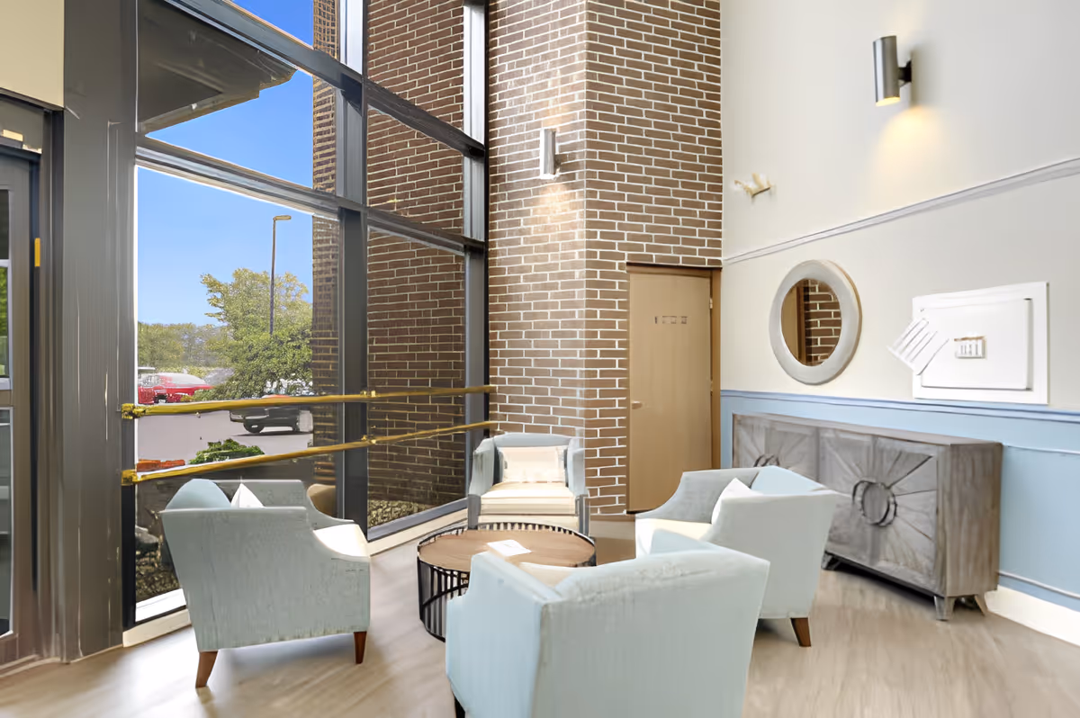 Sunlit seating area with light blue armchairs around a round table by floor-to-ceiling windows and a brick column.