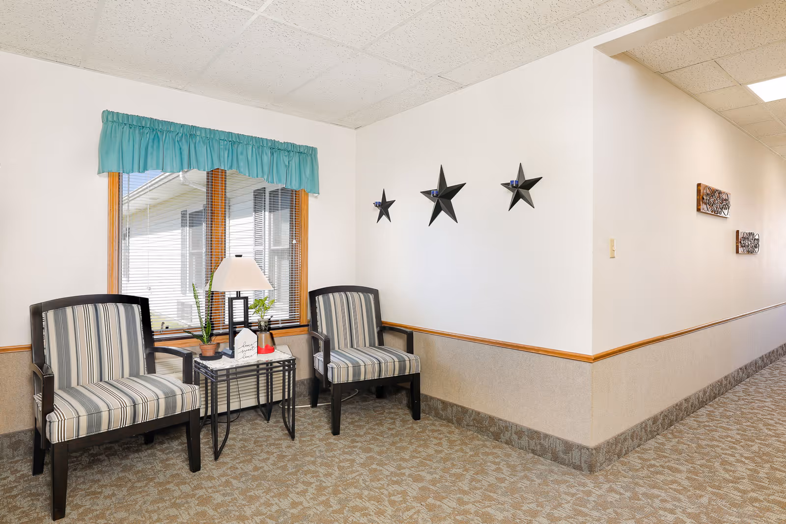 A small seating area in a hallway with two striped armchairs and a small table between them holding a lamp and two small plants. The wall behind the chairs has three black star-shaped candle holders. A window with a teal valance is above the chairs, and the hallway extends to the right with decorative wall hangings.