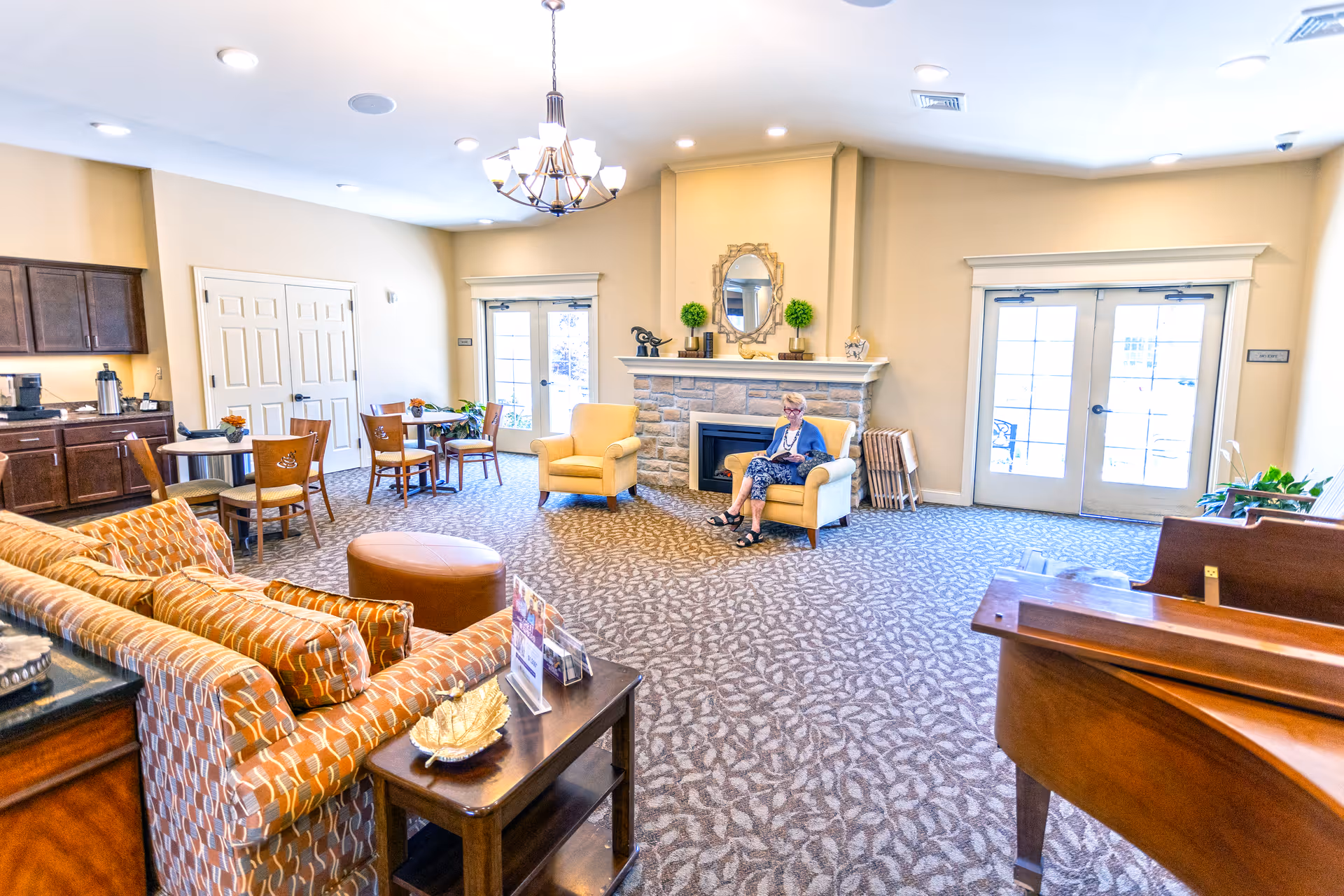 A bright communal living room with sofas, chairs, tables, a fireplace, and a person seated near the hearth.