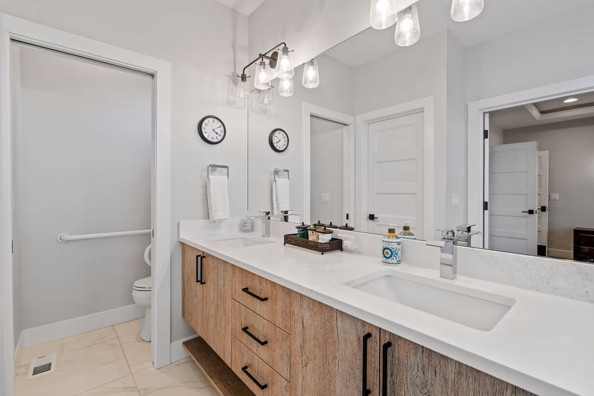 Modern bathroom with a double sink vanity featuring a white countertop and wooden cabinets. Above the sinks is a large mirror with a light fixture holding five glass shades. A clock and a towel holder with a white towel are mounted on the wall. To the left, there is a small room with a toilet and a grab bar. The bathroom has light-colored tiled flooring and white walls.