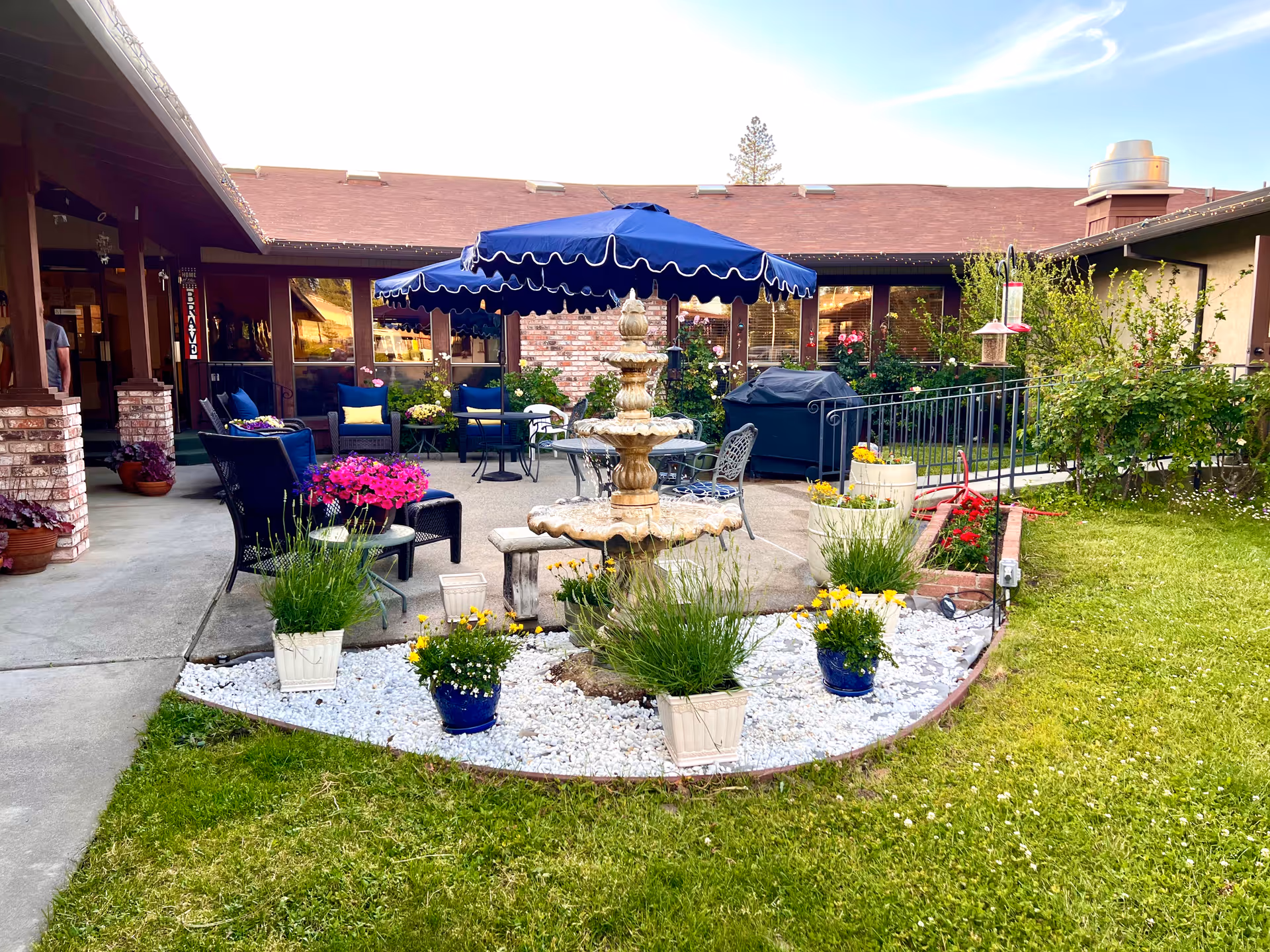 Outdoor patio area at Sierra View Senior Living featuring a central stone fountain surrounded by white gravel and potted plants with yellow and purple flowers. There are blue cushioned chairs and tables under large blue umbrellas, with a brick building and greenery in the background.