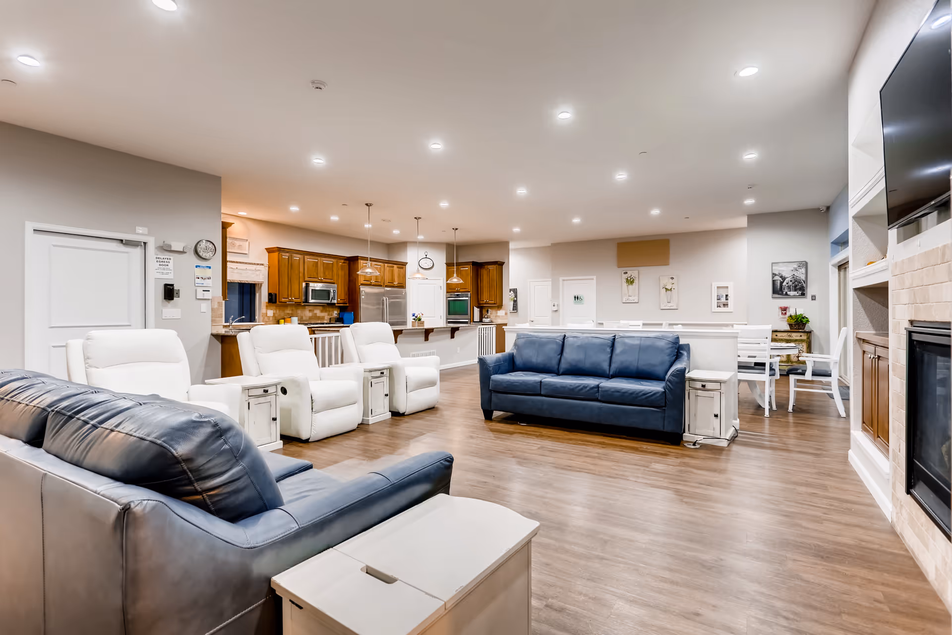 A spacious and well-lit common area in an assisted living facility featuring two blue leather sofas, three white recliner chairs, a fireplace with a mounted TV above it, and a kitchen area with wooden cabinets and stainless steel appliances in the background. There is also a dining area with white chairs and tables, wooden flooring, and recessed ceiling lights.