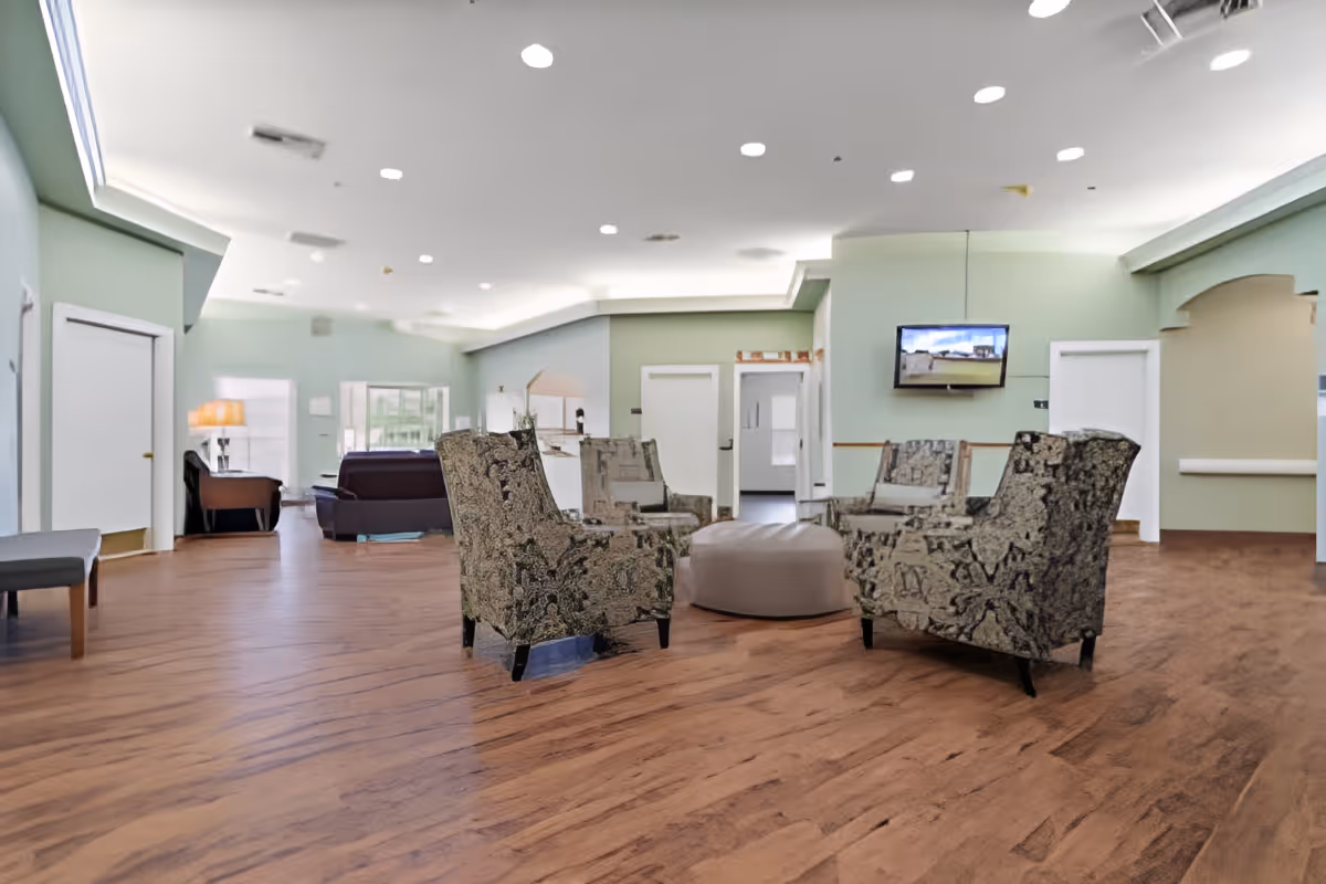 A spacious and well-lit common area with wooden flooring, several patterned armchairs arranged around a round ottoman, a wall-mounted TV, and multiple doorways leading to other rooms. The walls are painted in light green and beige tones, and the ceiling has recessed lighting.