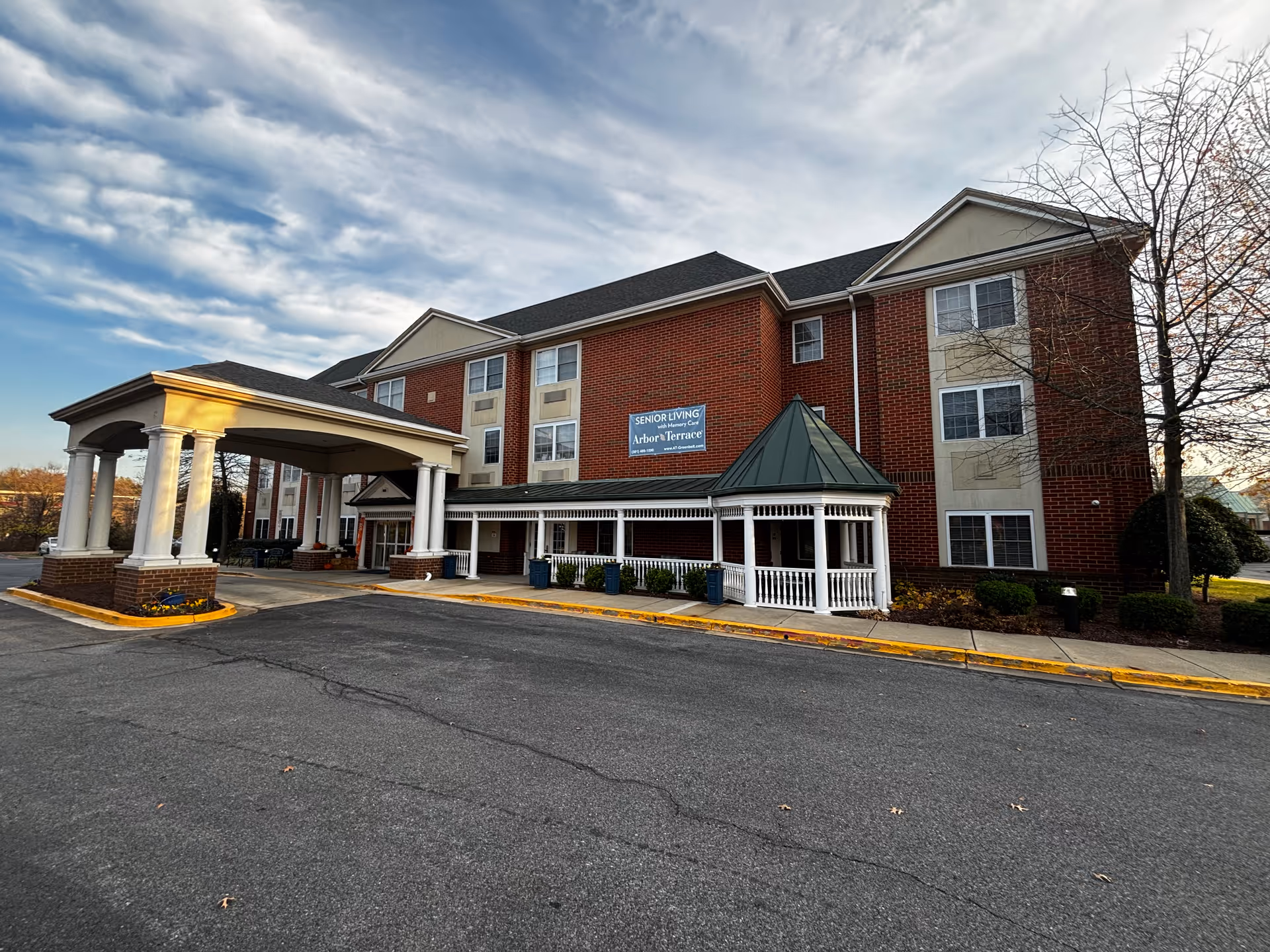 Exterior view of a three-story senior living facility with red brick and beige accents. The building has multiple windows and a covered entrance supported by white columns. There is a sign on the building that reads 'Senior Living with Memory Care Arbor Terrace'. The sky is partly cloudy.