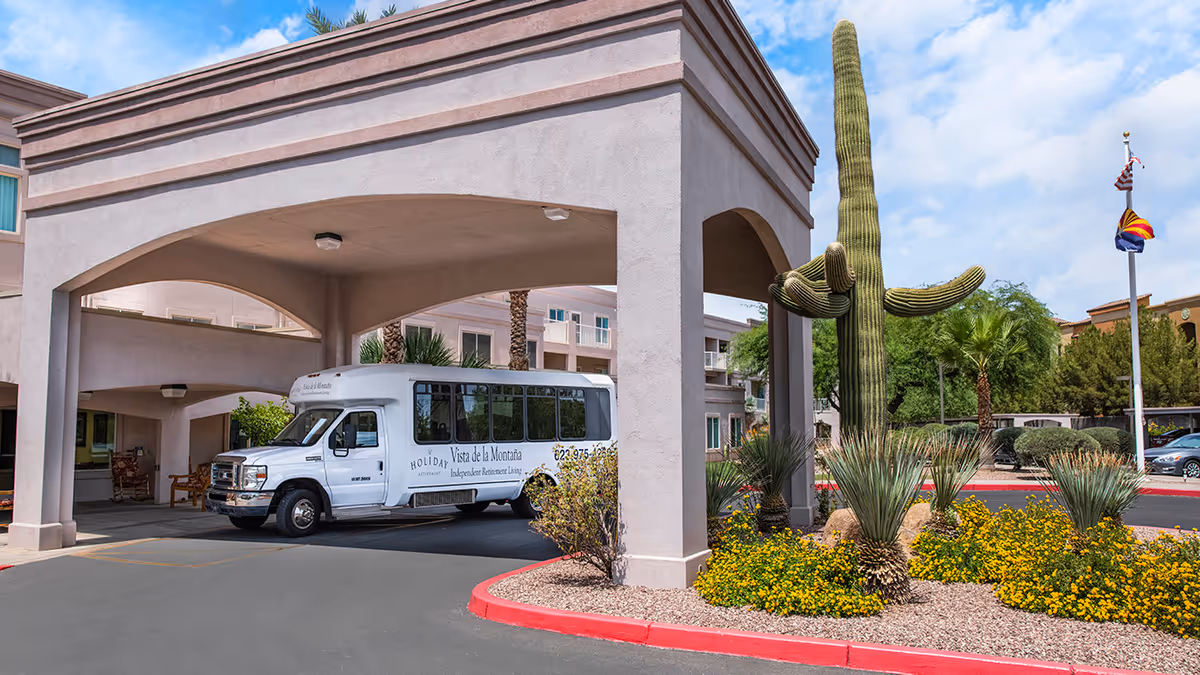 Entrance area of Holiday Vista de la Montana senior living facility with a white shuttle bus parked under a covered driveway. There is a large cactus and desert landscaping with yellow flowers near the curb. The building exterior is beige with multiple windows and palm trees in the background under a partly cloudy sky.