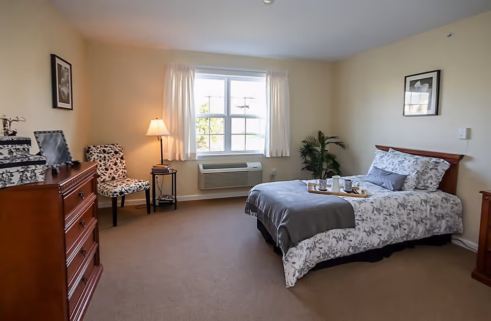 A cozy bedroom with a single bed made with floral patterned bedding and a gray throw blanket. A wooden dresser is on the left side, and a patterned chair with a small side table and lamp is placed near a window with white curtains. A green plant is in the corner, and two framed pictures hang on the beige walls.
