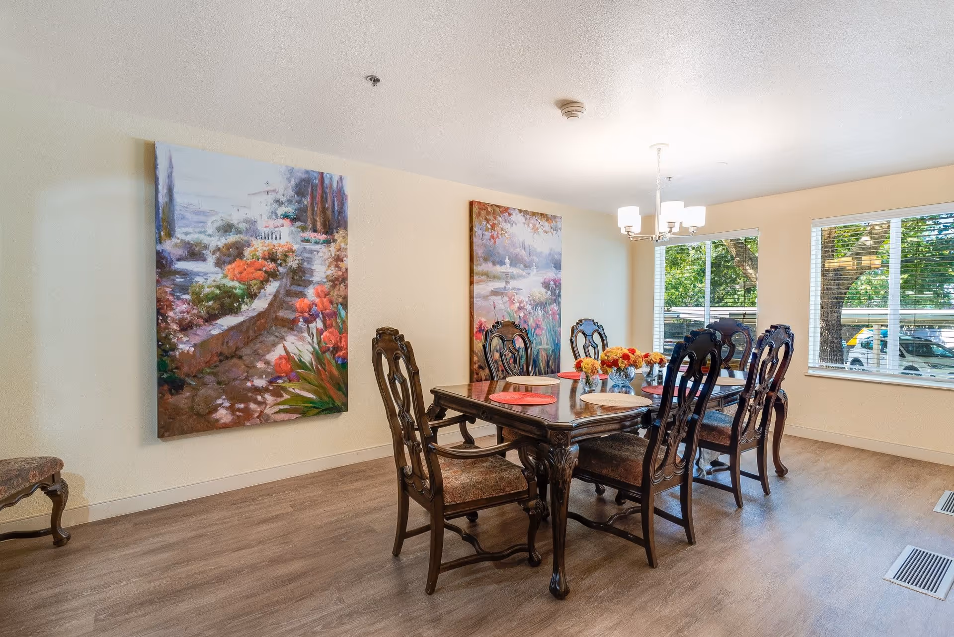 A dining room with a dark wooden table and six matching chairs with upholstered seats. The table is set with placemats and floral centerpieces. Two large colorful paintings of garden scenes hang on the light-colored walls. Large windows with blinds let in natural light and show trees and parked cars outside. The floor is wood with a light finish.