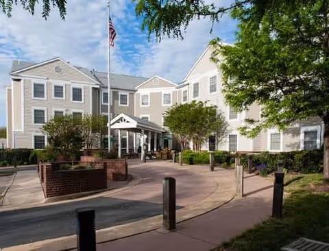 Exterior view of a three-story beige senior living facility building with white trim, an American flag on a flagpole in front, surrounded by trees and shrubs, with a curved driveway and walkway leading to the entrance.