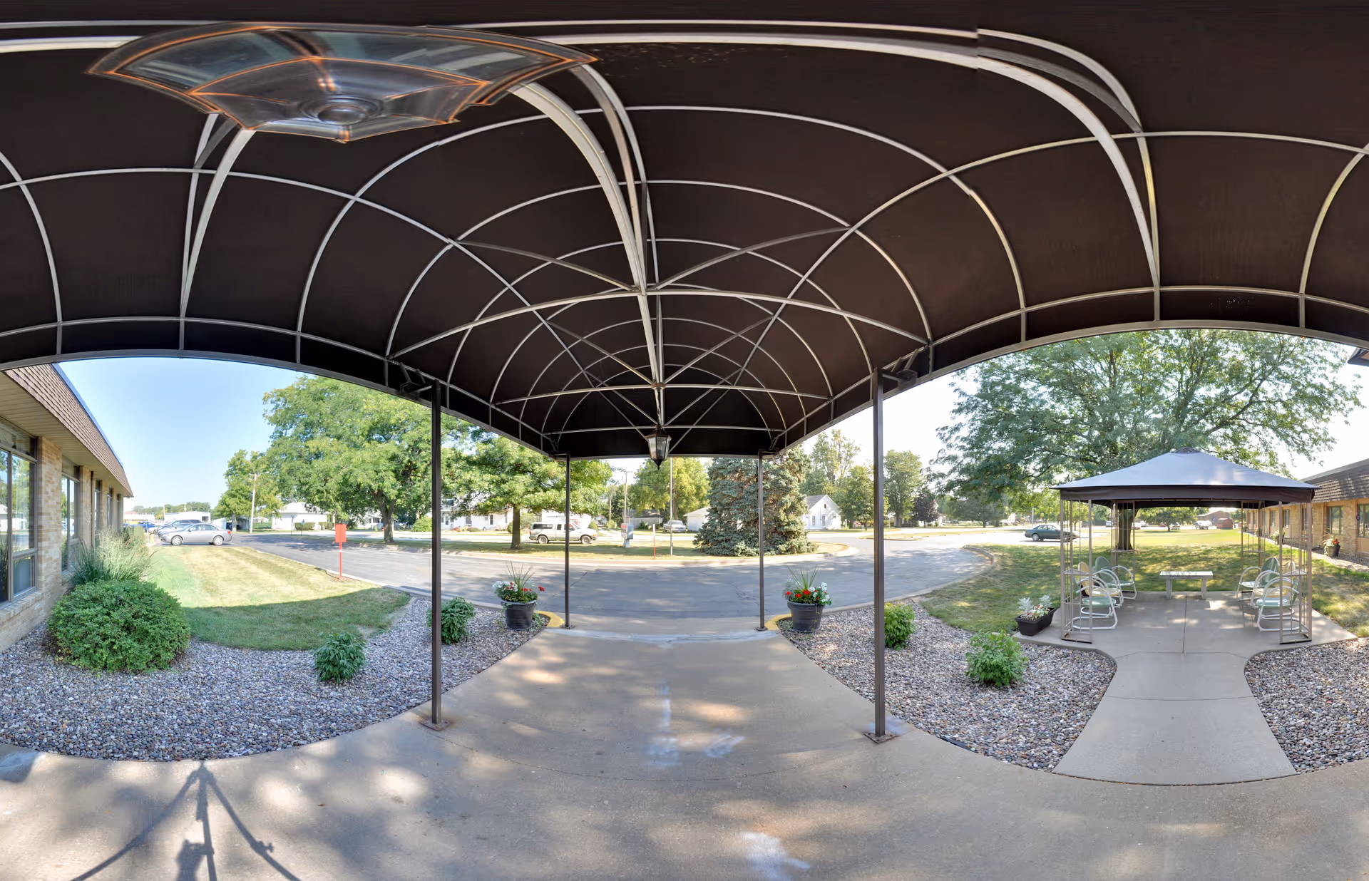 Covered entrance walkway with a metal canopy leading to a circular driveway, landscaped rock beds, and a gazebo to the right at the facility's front.