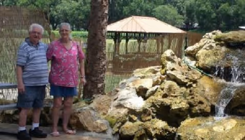 An elderly man and woman standing outdoors near a rock waterfall feature with a wooden gazebo and green trees in the background.
