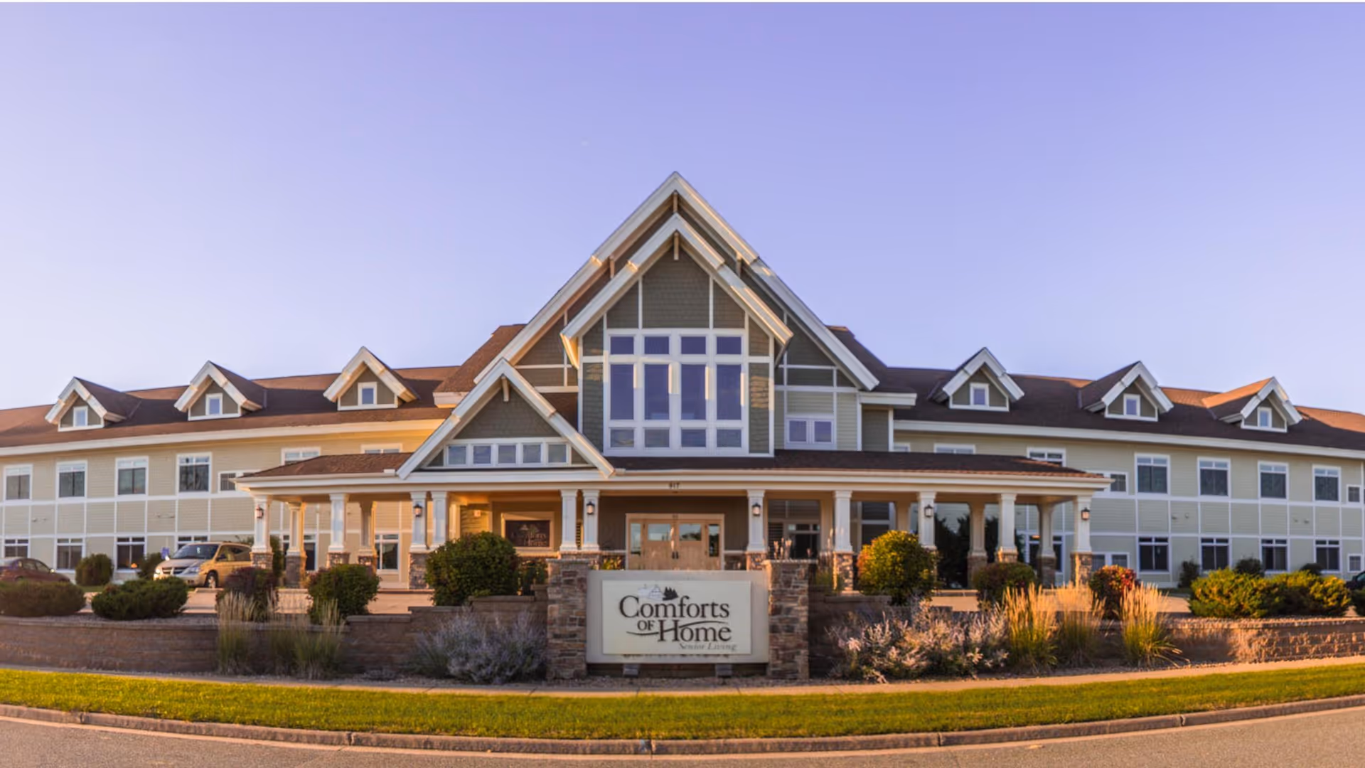 Front exterior of a two-story assisted living building with a landscaped entrance and a sign reading "Comforts of Home".