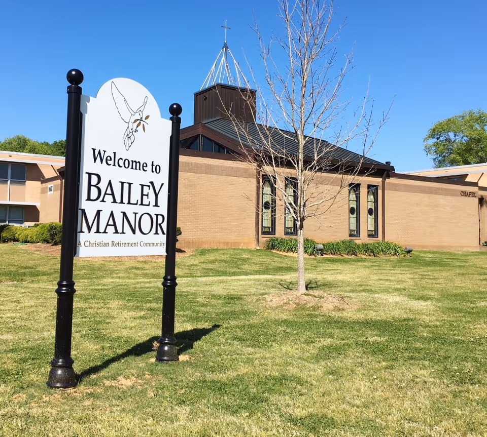 Outdoor view of a retirement community with a sign that reads 'Welcome to Bailey Manor A Christian Retirement Community' on a grassy lawn. Behind the sign is a brick building with stained glass windows and a small tree in front.