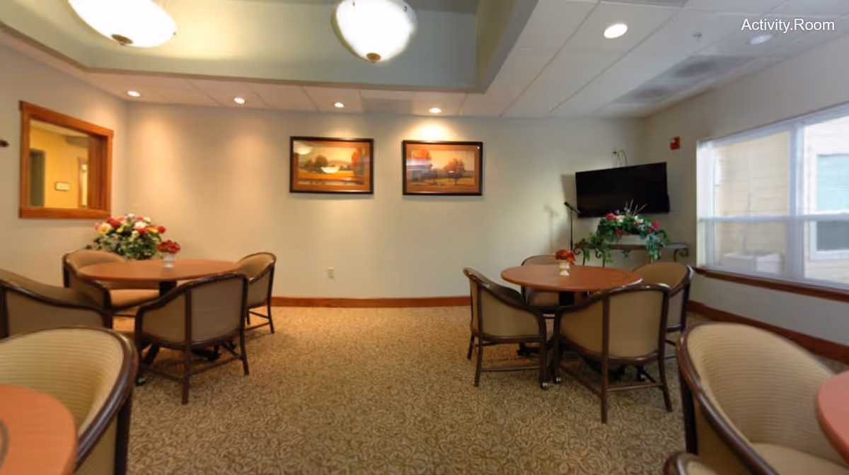 A well-lit activity room with round tables and cushioned chairs arranged around them. The room has beige walls with two framed landscape paintings, a wall-mounted TV, a window letting in natural light, and floral decorations on the tables and a stand beneath the TV.