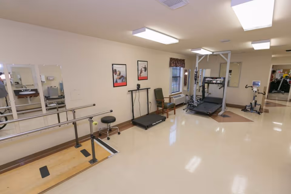 A rehabilitation room with parallel bars for walking exercises, a treadmill, an exercise bike, a chair, and mirrors on the wall. The room has beige walls, bright ceiling lights, and a window with a valance.