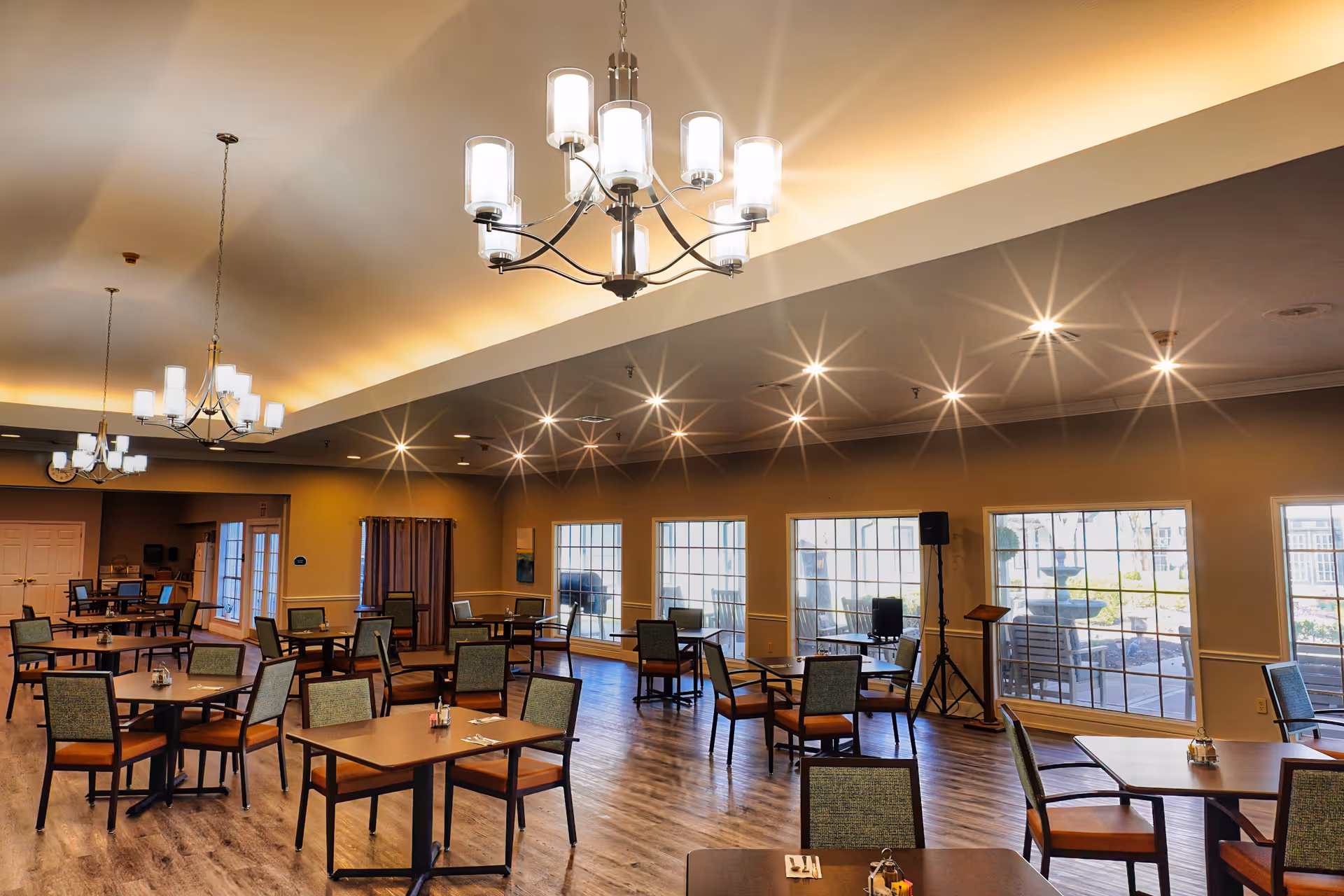 A spacious dining room with multiple square wooden tables and cushioned chairs arranged neatly. The room has large windows letting in natural light, wooden flooring, and modern chandeliers hanging from the ceiling. The walls are painted in a warm beige tone, and there is a speaker and podium near the windows.