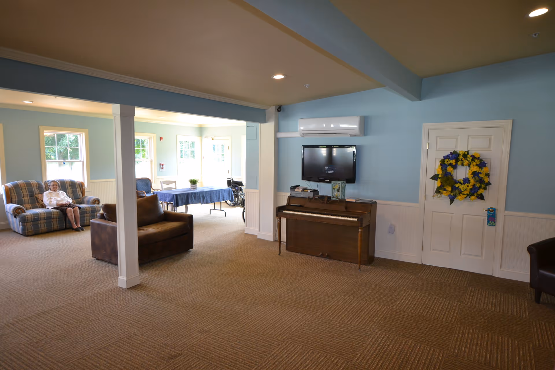 A spacious living area in an assisted living facility with light blue walls and beige carpet. The room features a plaid armchair with an elderly woman sitting on it, a brown leather armchair, a piano with a flat-screen TV mounted above it, and a door decorated with a yellow and blue flower wreath. In the background, there is a table covered with a blue tablecloth and a wheelchair near the windows.