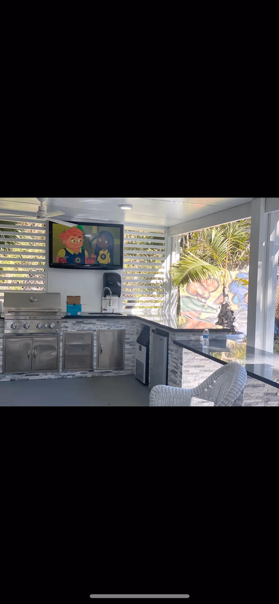 Outdoor covered kitchen area with stainless steel grill, sink, refrigerator, and countertop. A flat-screen TV mounted on the wall shows an animated cartoon. There is a white wicker chair and tropical plants visible outside.