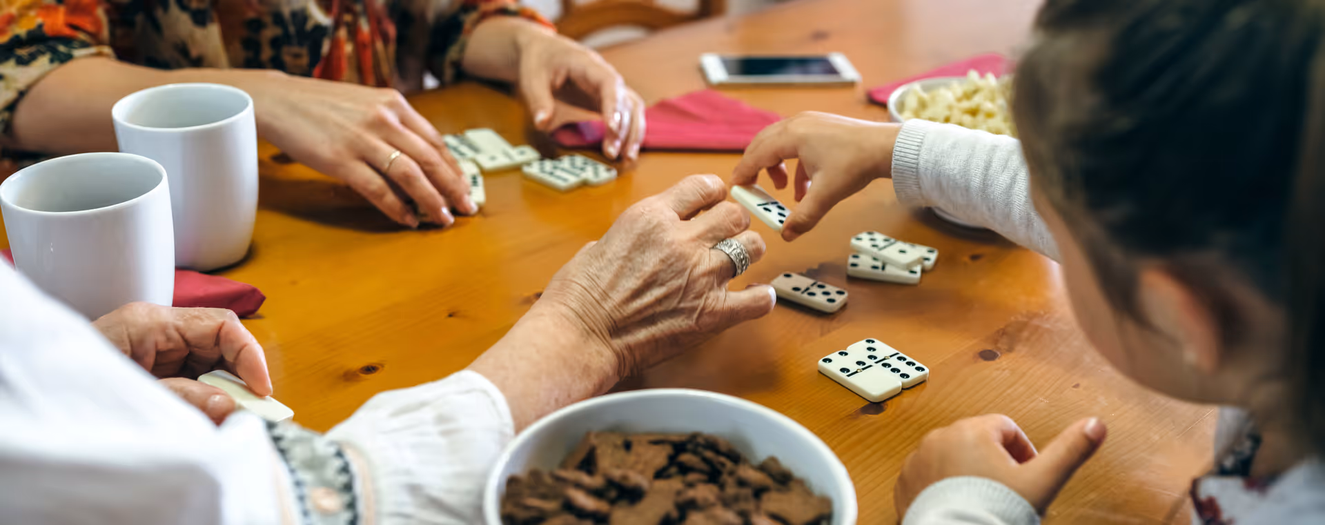 Several people sitting around a wooden table playing dominoes, with bowls of snacks and two white mugs on the table.